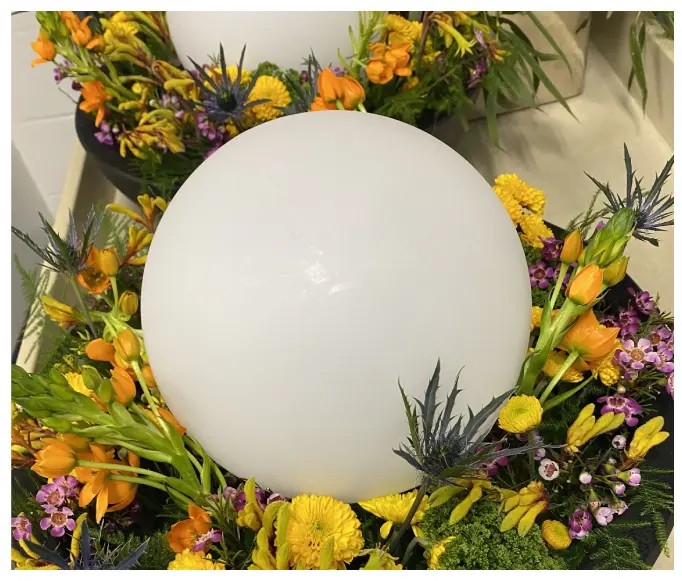 A white spherical lamp is surrounded by a vibrant arrangement of yellow, orange, and purple flowers with green foliage.